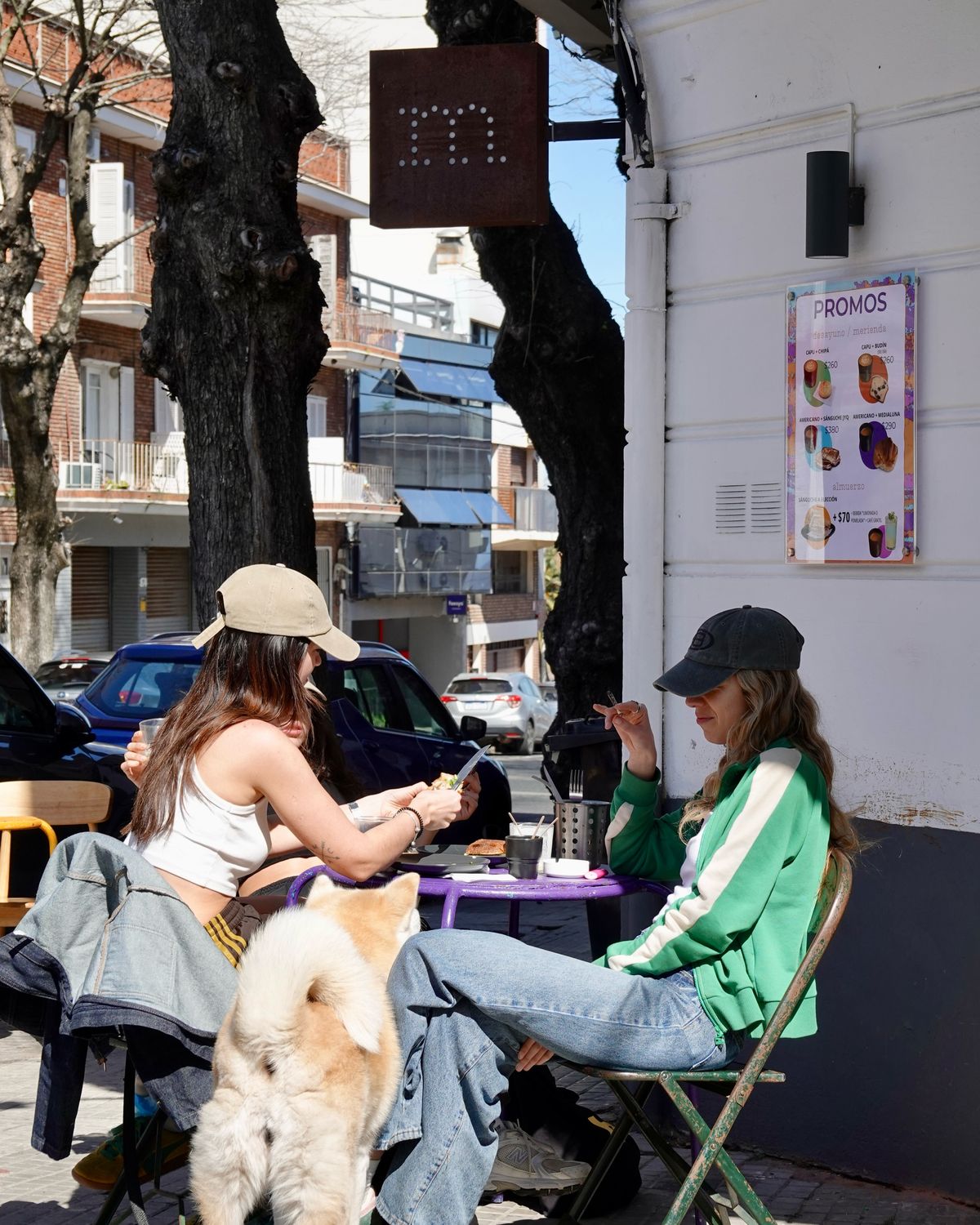 Dos amigas en la mesita violeta de la vereda, con un perro y el cartel de Mirador